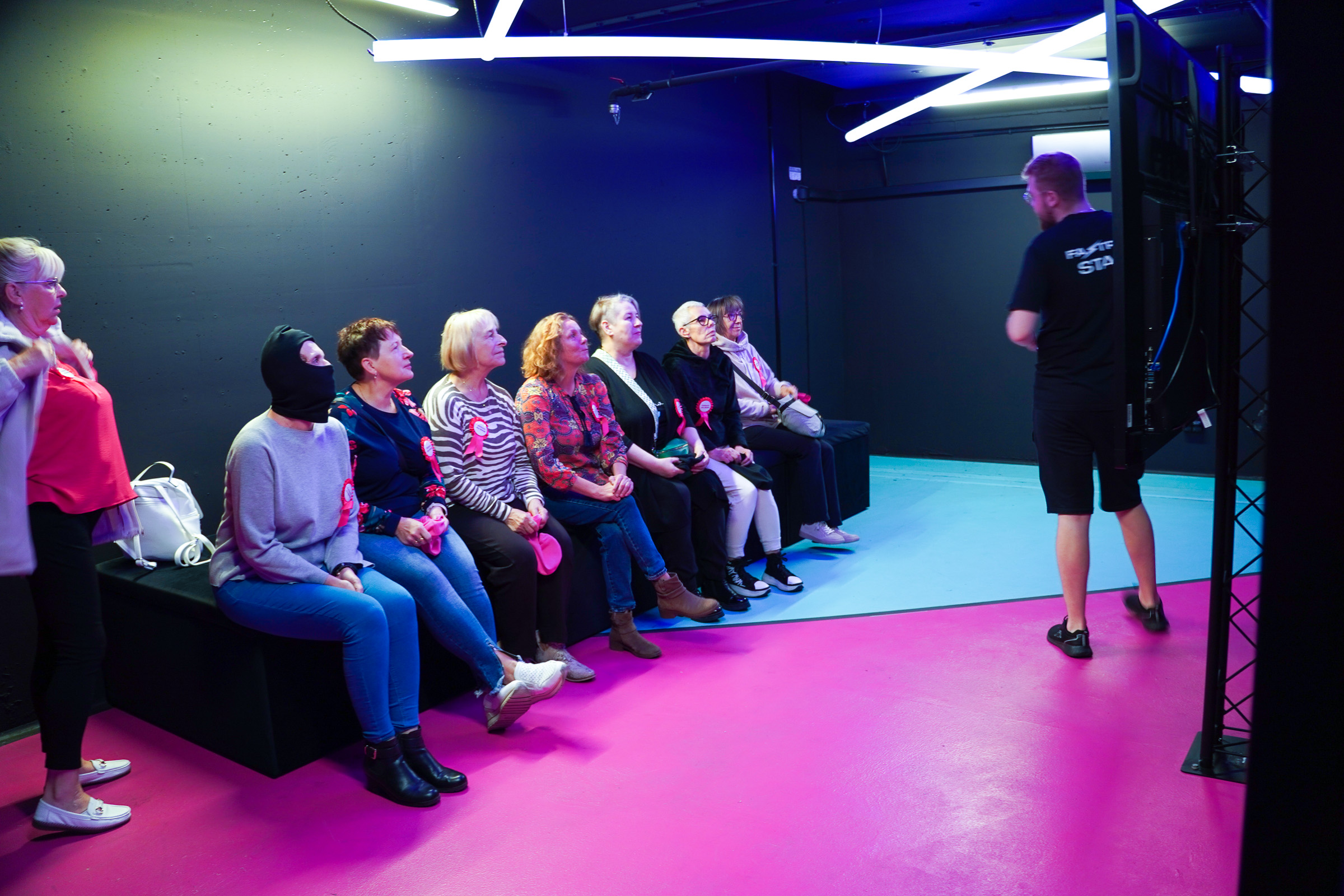 Kilkanaście kobiet siedzi w oświetlonym kolorowym światłem pomieszczeniu toru Fast Track, słuchając instruktora tłumaczącego zasady jazdy gokartami.